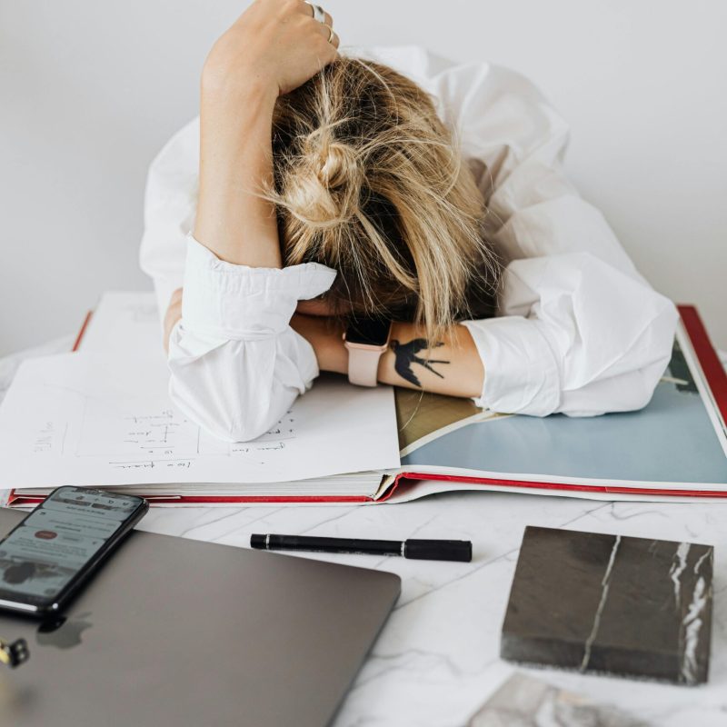 Overwhelmed woman resting head on desk with laptop and papers in home office.